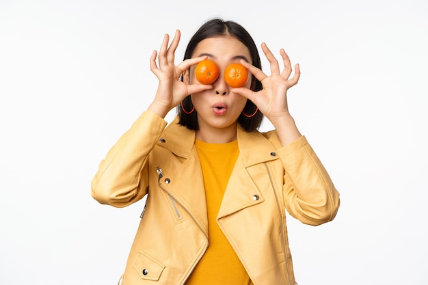 Funny asian girl holding tangerine on eyes and smiling making playful grimaces standing over white background