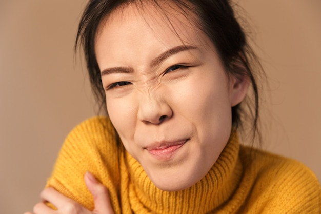 Funny asian woman wearing sweater grimacing while posing at front in studio isolated over beige wall