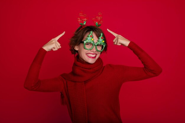 Funny attractive young lady in Christmas reindeer horns and glasses is posing and enjoying on party on red background Merry christmas Happy new year Holidays and celebration in winter