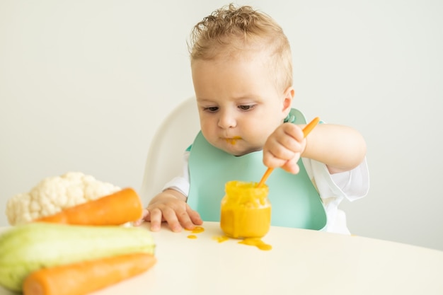 Funny baby boy in bib eating vegetable puree with spoon sitting in child chair. Child learn to eat by himself.