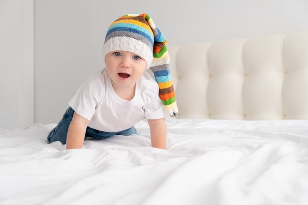 Funny baby boy in funny colorful hat smiling and lying on a white bedding at home.