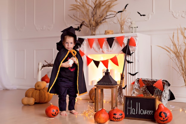 Funny baby boy in halloween witch costume with pumpkin jack and broom indoors