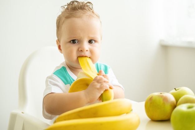 Funny baby boy sitting at the table in child chair eating banana on white kitchen.