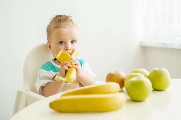 Забавный мальчик, сидящий за столом в детском кресле, ест банан на белой кухне.
