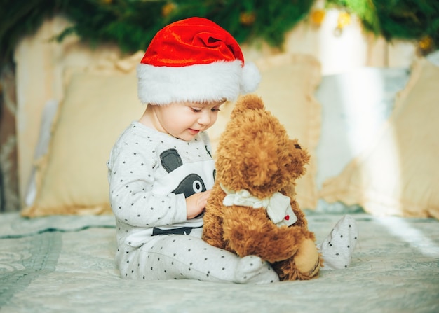 Funny baby boy wearing santa hat and pajama sitting on the bed with favorite toy. First Christmas. New Year's holidays.