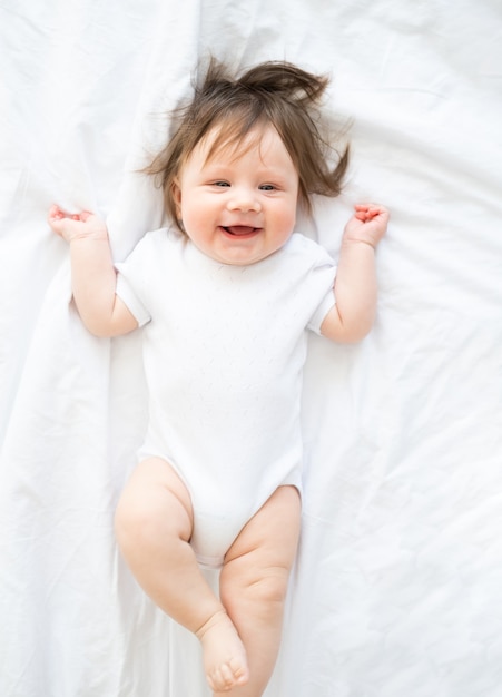 Funny baby boy in white bodysuit smiling and lying on a white bedding at home. top view.