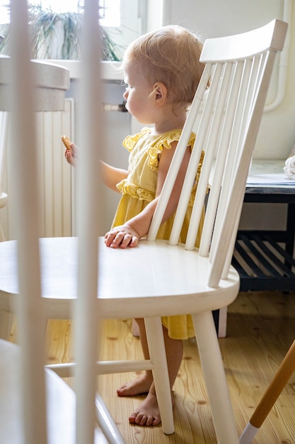 Foto bambina divertente che mangia biscotti fatti in casa in piedi in una cucina moderna vicino a una sedia e un tavolo di legno bianco