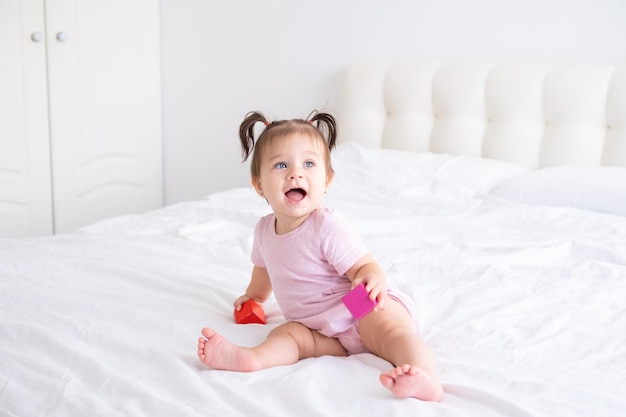 ベッドの上の子供の木製キューブおもちゃで遊ぶピンクのボディー スーツで面白い女の赤ちゃん