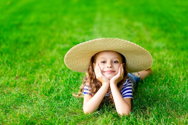 Funny baby girl in summer on the lawn with a big straw hat on the green grass having fun and rejoicing, space for text