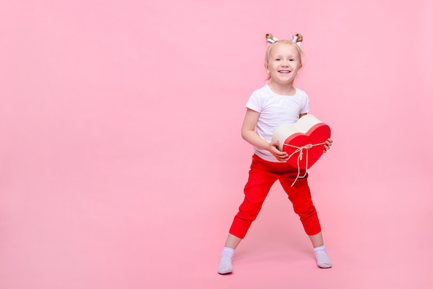 Funny baby girl in a white t-shirt and red pants with a heart-shaped box on a pink background. Children's portrait with space for text.