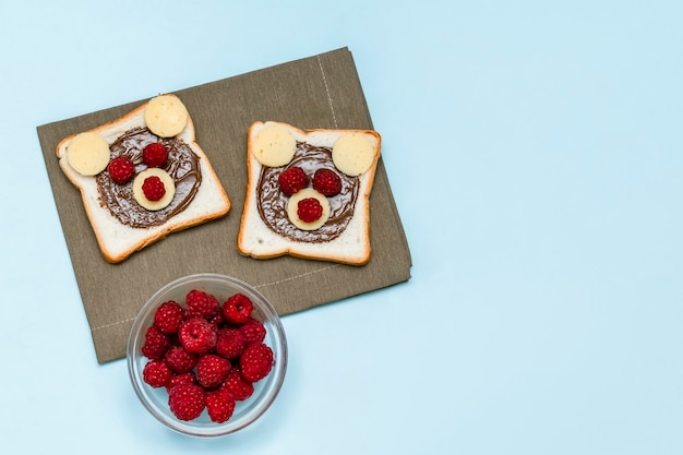 Funny bear face sandwich toast bread with peanut butter, cheese and raspberry on napkin blue background. Kids child sweet dessert breakfast lunch food close up