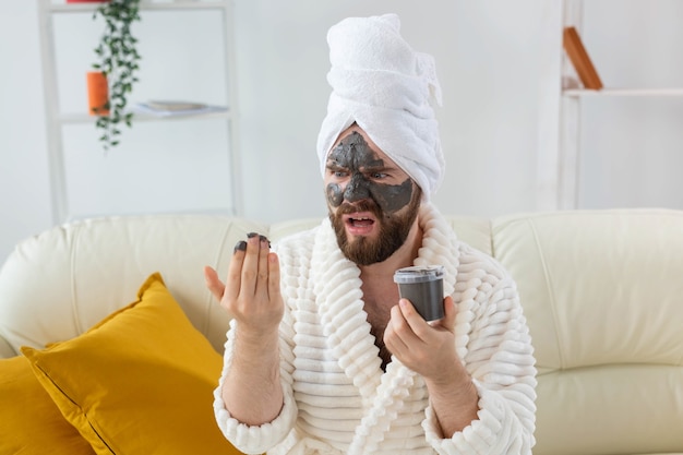 Funny bearded man having fun with a cosmetic mask on his face made from black clay men skin care