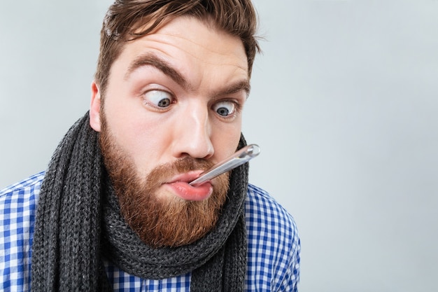 Фото Забавный бородатый молодой человек в шарфе с термометром во рту над белой стеной