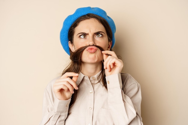 Funny beautiful girl fooling around making moustache out of hair under lips squinting and grimacing ...