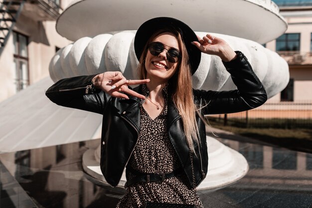Фото Смешная красивая девушка с улыбкой в модном платье с кожаной курткой, шляпой и солнцезащитными очками показывает знак мира и сидит в городе