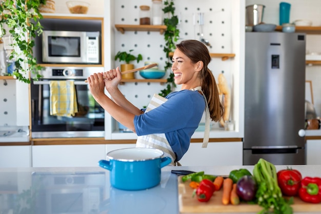 Funny beautiful woman singing into spatula cooking in modern kitchen holding spatula as microphone dancing listening to music playful girl having fun with kitchenware preparing food