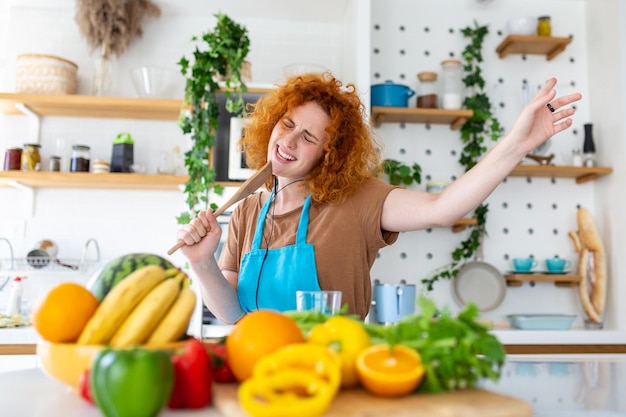 Funny beautiful woman singing into spatula cooking in modern kitchen holding spatula as microphone dancing listening to music playful girl having fun with kitchenware preparing food