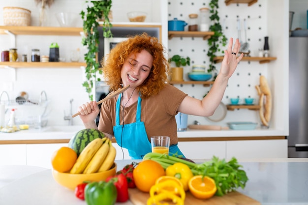 Funny beautiful woman singing into spatula cooking in modern kitchen holding spatula as microphone dancing listening to music playful girl having fun with kitchenware preparing food