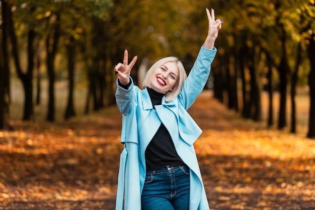 Funny beautiful young girl with a white smile in a fashion blue coat shows a peace sign in the park with autumn foliage