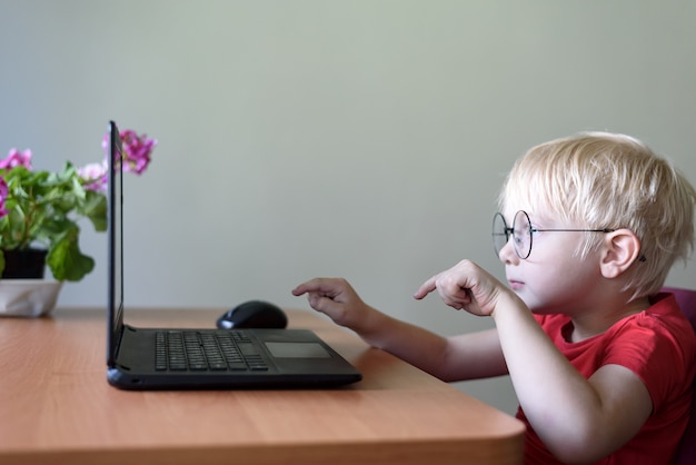 Funny blond boy in glasses sits at a laptop