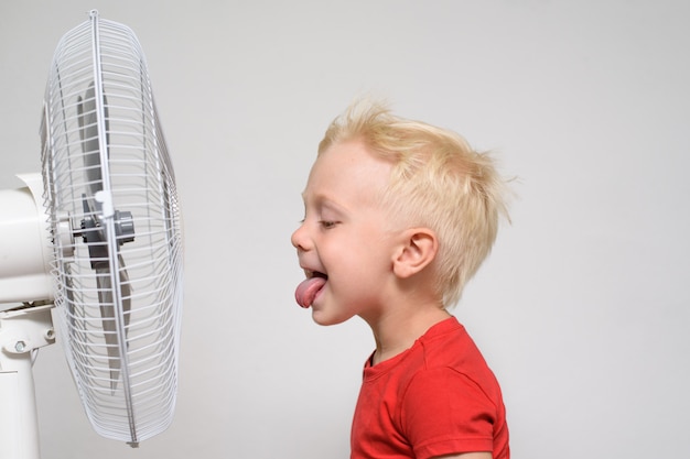 Funny blond boy in a red T-shirt near the fan with his tongue sticking out. Enjoy cool air. Summer concept