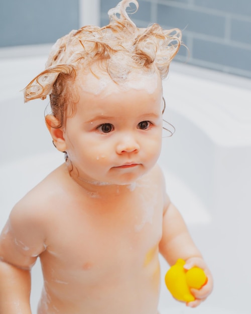 Funny blond kid boy having fun with water by taking bath in bathtub washing adorable baby in bathroo