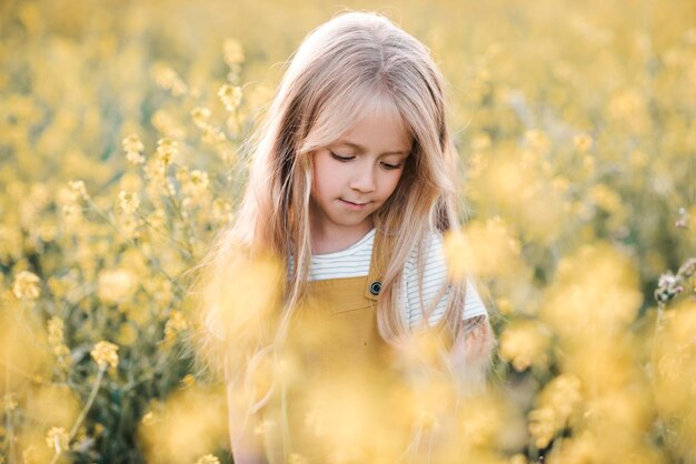 自然の背景の上に菜の花の花の咲く牧草地でポーズをとって面白いブロンドの子供の女の子