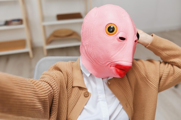 Funny blonde lady in pink fish mask warm sweater doing selfie\
use phone sitting in armchair at modern home interior pause from\
work take a break social media in free time concept wide angle