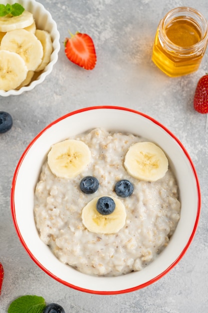 Funny bowl with oat porridge with bear faces made of fruits and berries on a gray concrete background. Food for kids idea, top view, copy space.