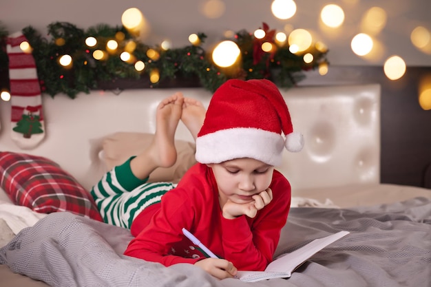 Funny boy kid in Santa hat writing letter to Santa Claus lie on a bed