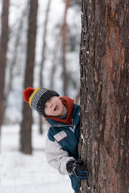 Funny boy peeks out from behind tree Child in knitted hat walks through winter snowkept park