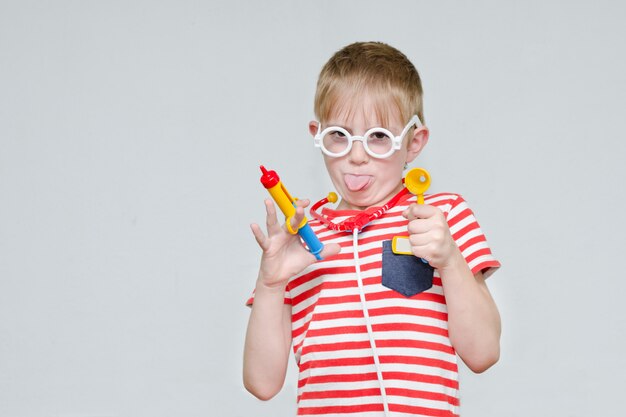 Funny boy playing doctor. Toy syringe, glasses and phonendoscope. Portrait