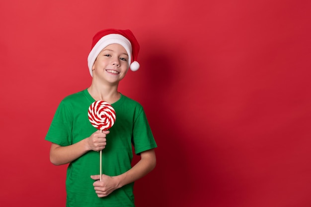 funny boy in santa hat and green tshirt holding lollipop on red background