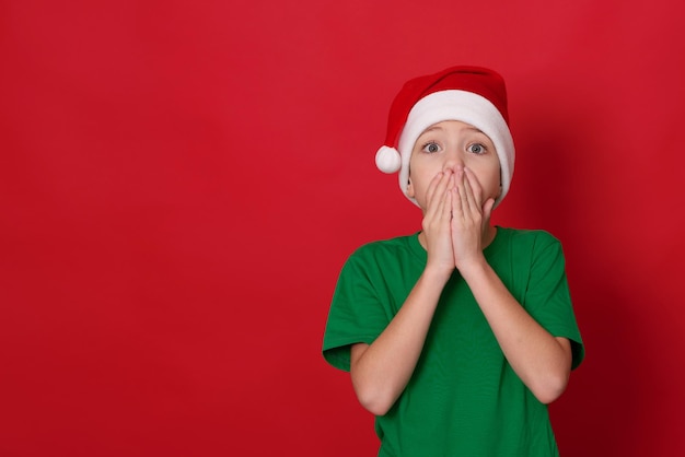 funny boy in santa hat and green tshirt is making faces showing surprise