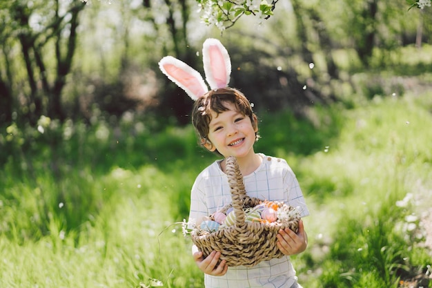 Funny boy with eggs basket and bunny ears on Easter egg hunt in sunny spring garden