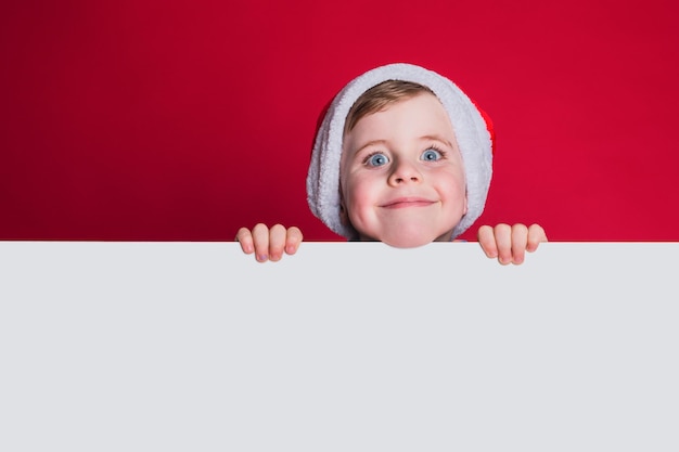 Photo funny boy with santa claus hat on red background, free space for text.
