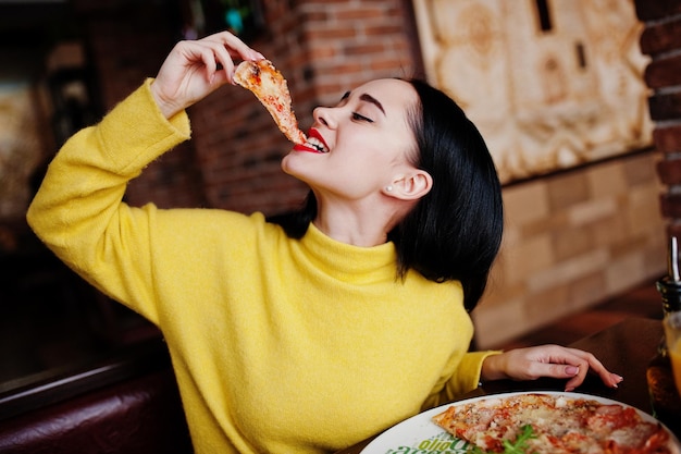 Funny brunette girl in yellow sweater eating pizza at restaurant