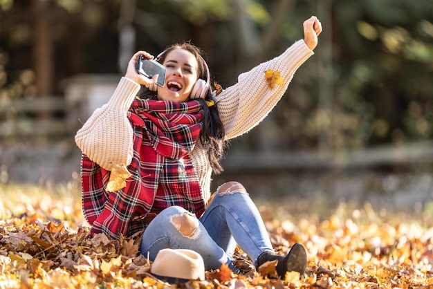 Funny brunette in stylish autumn clothes holding phone as a mic singing in the autumn leaves listening to music with headphones