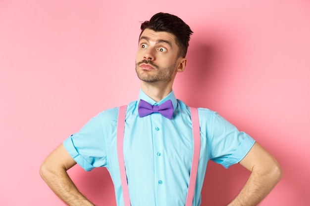 Funny caucasian man with moustache and bow-tie, standing confident with hands spread sideways, looking left at logo proud, standing on pink.