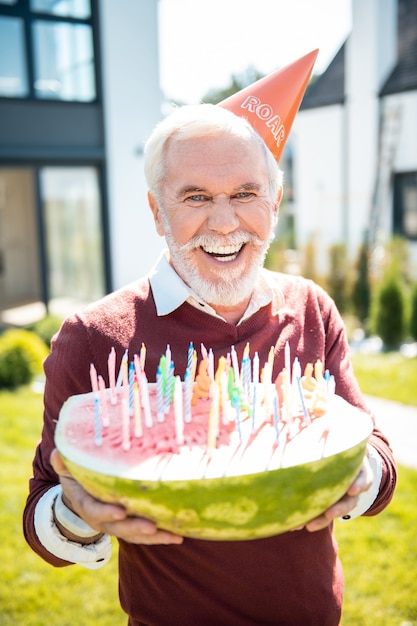Funny celebration. Cheerful pensioner wearing paper hat while demonstrating his watermelon with candles