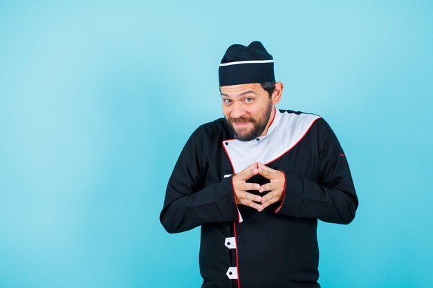 Funny chef is looking at camera by holding hands together on blue background