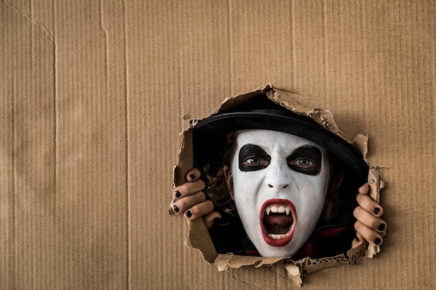 Photo funny child dressed halloween costume looking through hole on cardboard. kid painted terrible vampire. autumn holiday concept