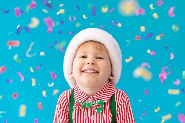 Funny child dressed Santa Claus hat against blue background. Happy kid having fun at home. Christmas holiday concept