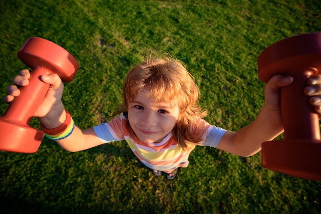 Funny child exercise with dumbbells on grass outside strong little boy children health concept outsi...