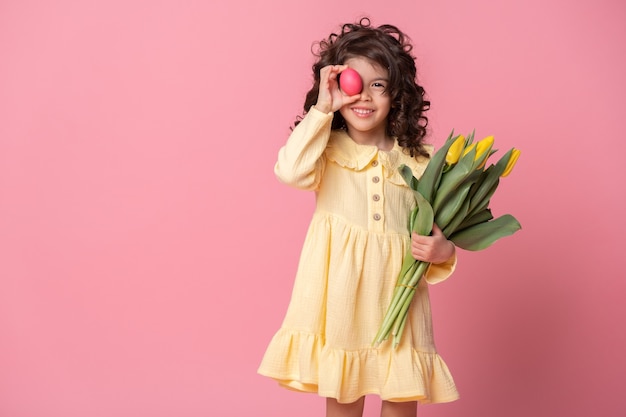 Funny child girl holding tulips and colorful Easter egg in front of her eye on pink background.