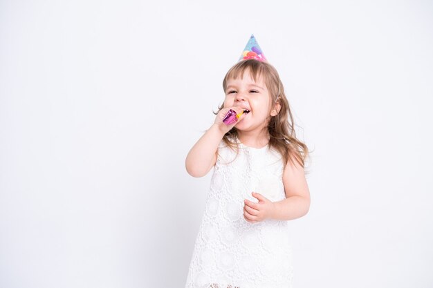 Funny child girl in white dress and birthday hat blowing in whistle on white surface.