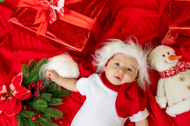 A funny child in a Santa costume lies on a red background among gifts