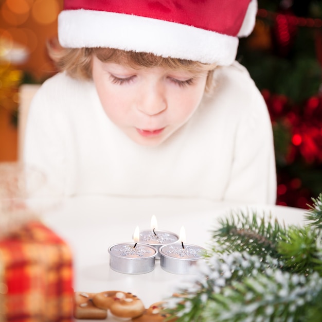 Funny child in Santa hat blowing out burning Christmas candles Focus on candles