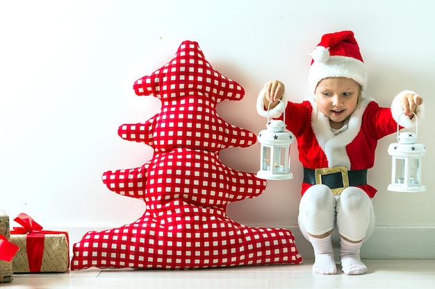 funny child in a suit of Santa Claus sitting near a red textile Christmas tree