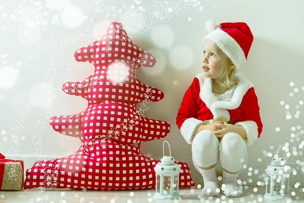 funny child in a suit of Santa Claus sitting near a red textile Christmas tree